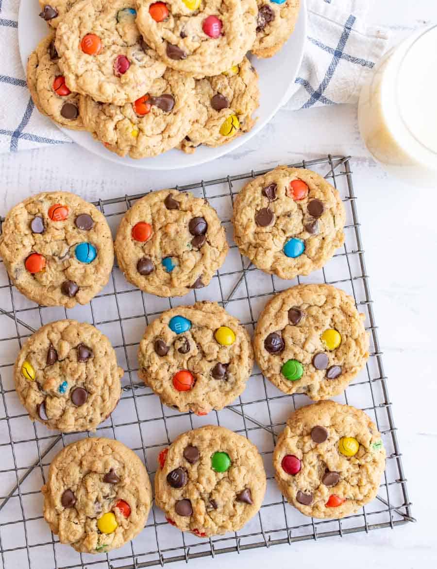 A cooling rack holds oatmeal cookies with colorful candy-coated chocolates and chocolate chips. More cookies are stacked on a plate nearby, with a glass of milk in the corner.