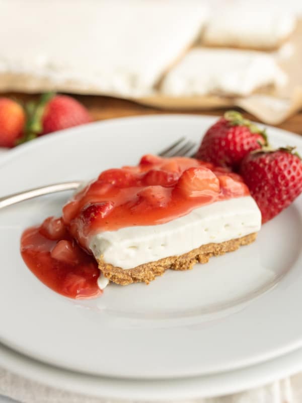 A slice of cheesecake with a graham cracker crust, topped with strawberry sauce, is served on a white plate next to two fresh strawberries and a fork. More dessert bars and strawberries are visible in the background.