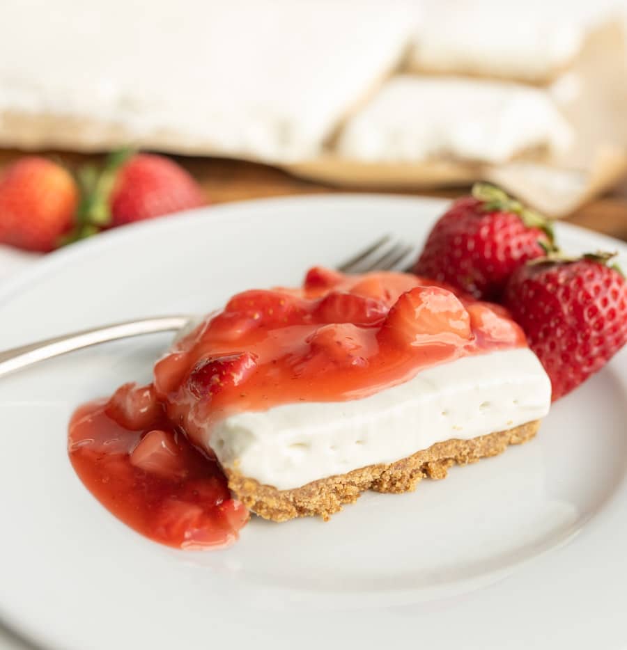 A slice of cheesecake with a graham cracker crust is topped with strawberry sauce and served on a white plate, accompanied by two fresh strawberries and a fork.