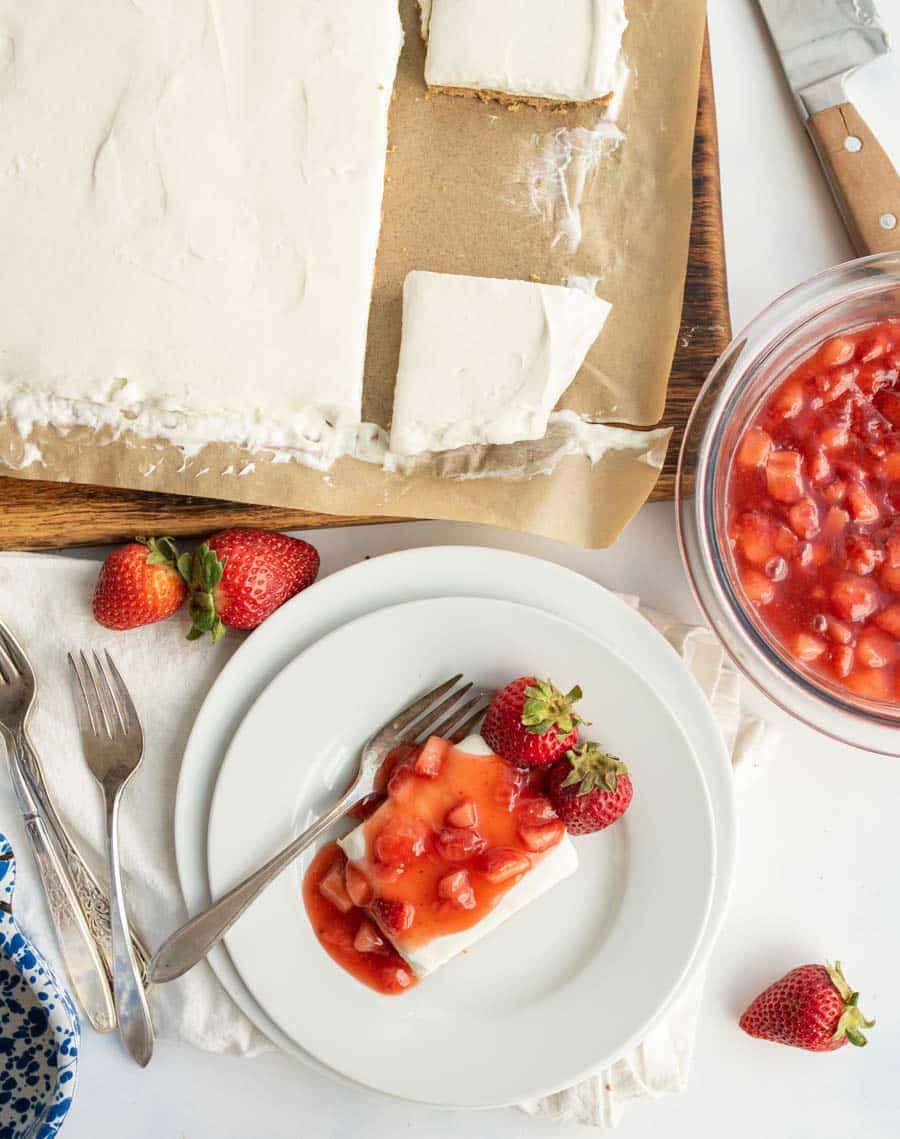 A white-frosted cake, partly sliced, sits on parchment paper. A plate holds a cake slice topped with strawberry sauce and fresh strawberries. A bowl of sauce, strawberries, forks, and a knife are nearby on a white surface.