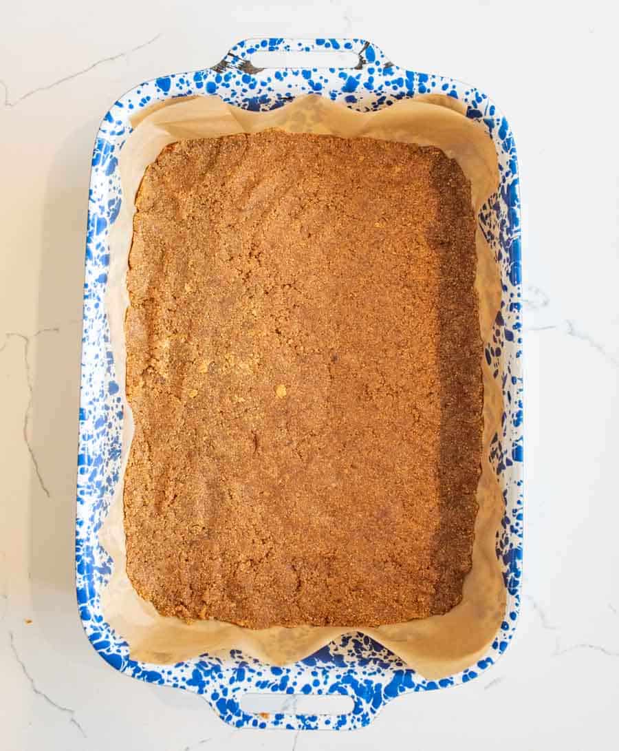 A baking dish lined with parchment paper holds a large, rectangular, golden-brown baked dessert with a crumbly texture, set on a white marble surface.