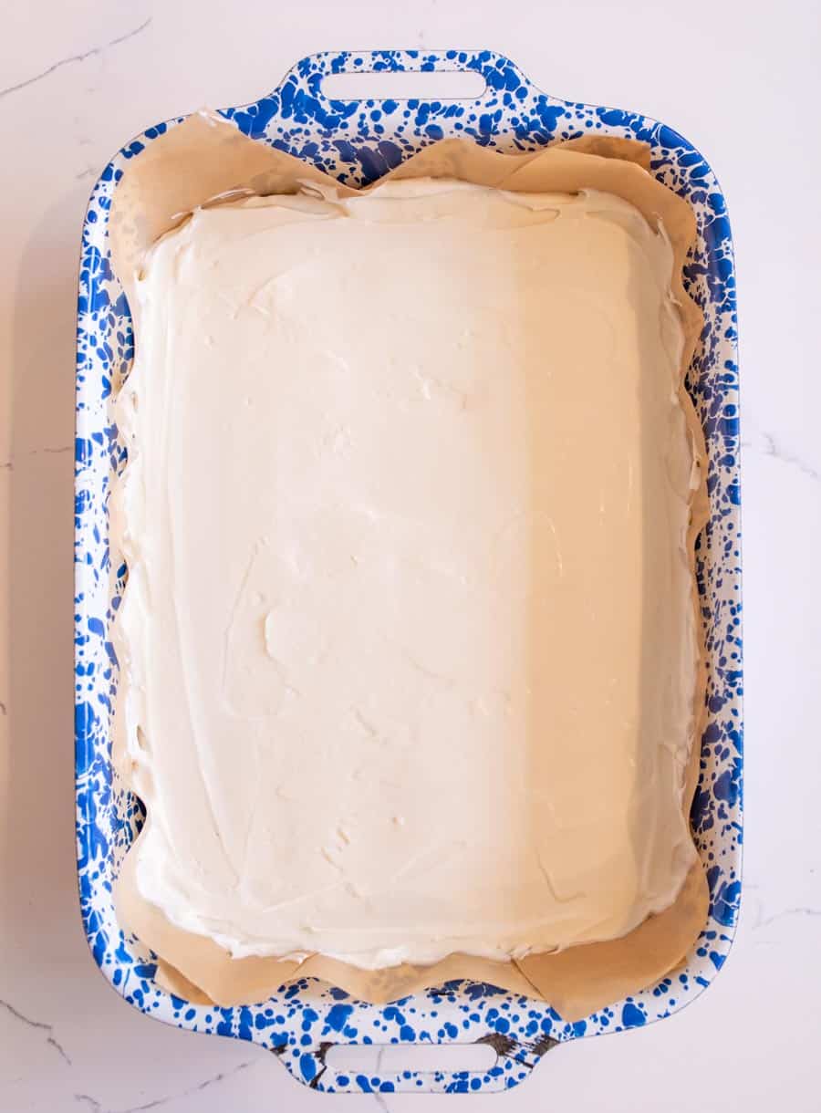 A rectangular cake with smooth white frosting sits in a blue and white speckled baking dish lined with parchment paper, on a white surface.