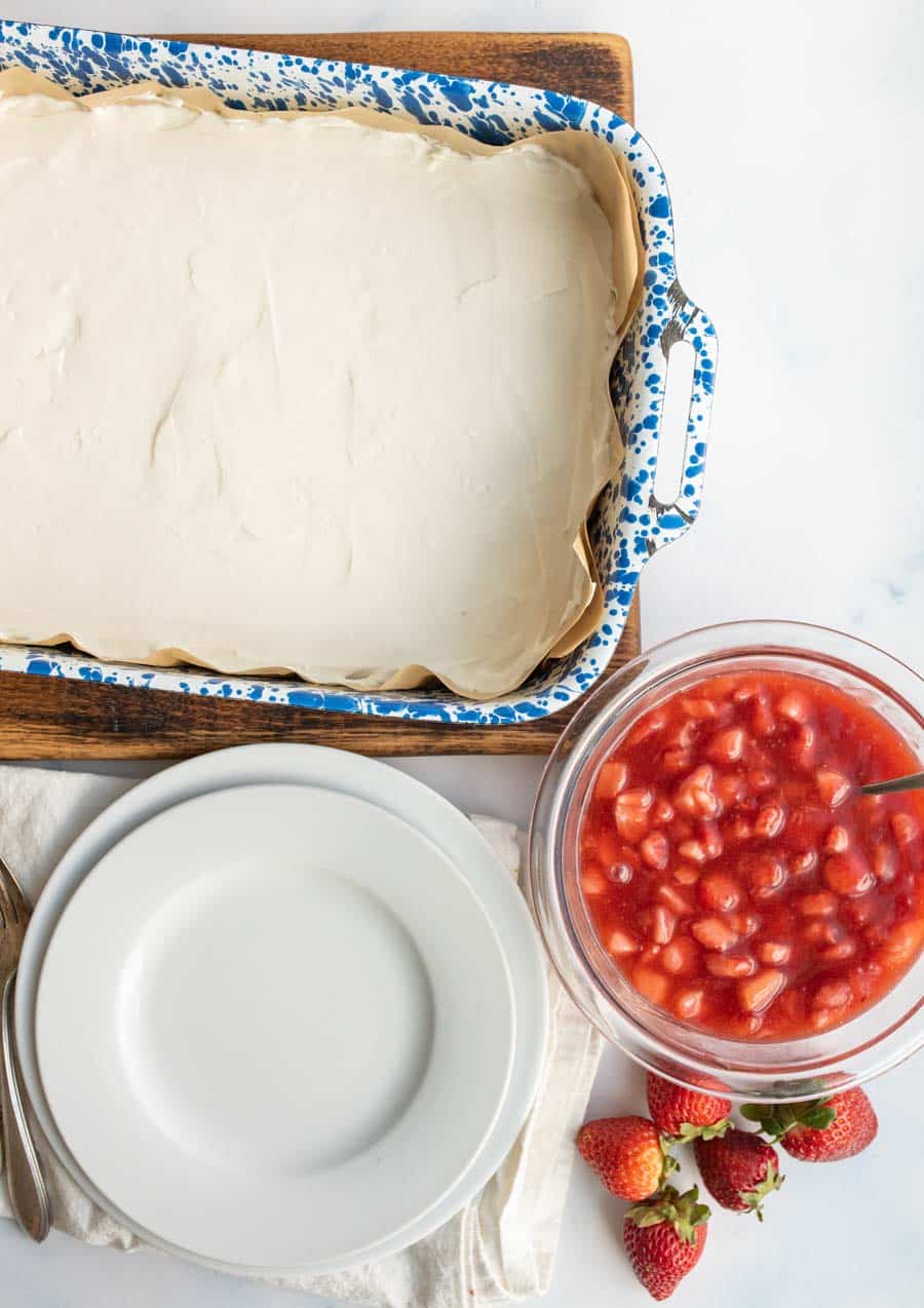 A blue speckled baking dish filled with a creamy white dessert sits beside a bowl of strawberry topping and a stack of white plates on a white surface, with fresh strawberries nearby.