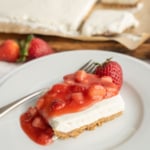 A slice of cheesecake with a graham cracker crust topped with strawberry sauce and a whole strawberry sits on a white plate with a fork; more cheesecake and strawberries are in the background.