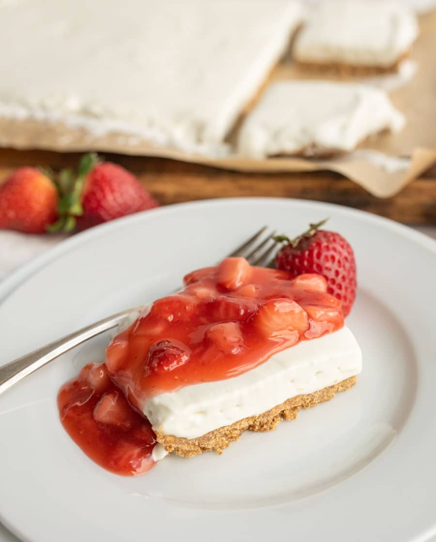 A slice of cheesecake with a graham cracker crust topped with strawberry sauce and a whole strawberry sits on a white plate with a fork; more cheesecake and strawberries are in the background.