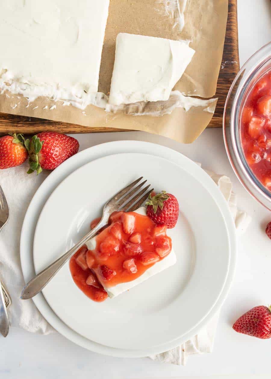 A slice of creamy white dessert topped with strawberry sauce and a whole strawberry sits on a white plate with a fork. Nearby are more strawberries, a glass bowl of strawberry sauce, and the remaining dessert on a wooden tray.