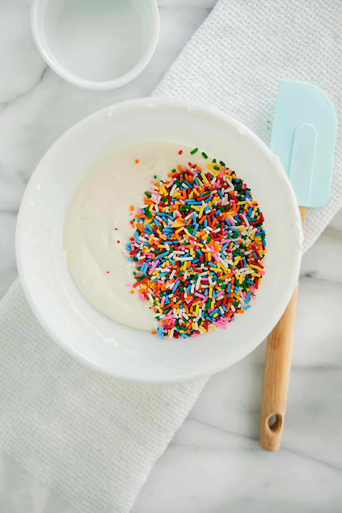 A white bowl with white cake batter and colorful rainbow sprinkles sits on a white textured cloth, next to a blue spatula and a small empty dish on a marble countertop.