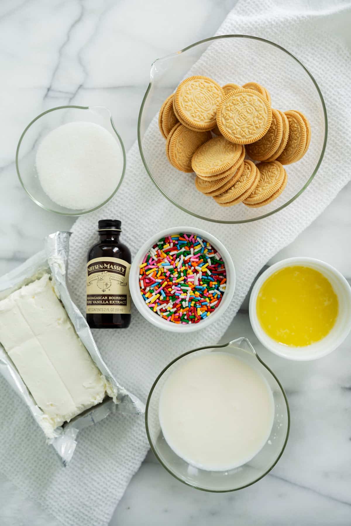 A flat lay of dessert ingredients on a white surface: golden sandwich cookies, white sugar, colorful sprinkles, cream cheese, vanilla extract, melted butter, and heavy cream in various bowls and containers.