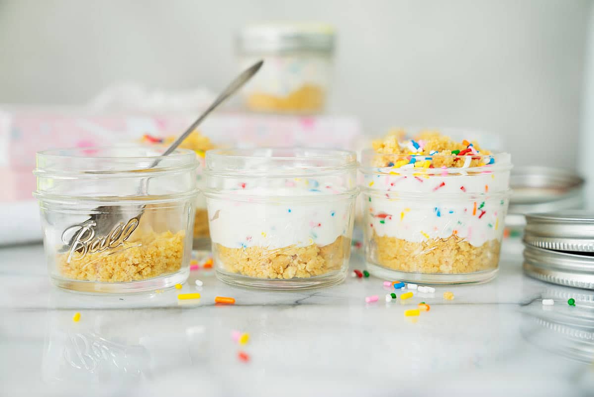 Three small glass jars are filled with layered dessert, featuring a crumbly crust, creamy filling, and colorful sprinkles on top. One jar is partially eaten, and loose sprinkles are scattered around.