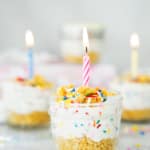 A small glass jar filled with layered cake and creamy frosting, topped with colorful sprinkles and a lit pink-striped candle. Similar jars with candles and sprinkles are blurred in the background.
