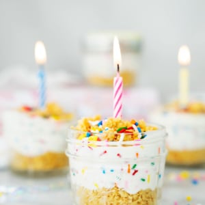 A small glass jar filled with layered cake and creamy frosting, topped with colorful sprinkles and a lit pink-striped candle. Similar jars with candles and sprinkles are blurred in the background.