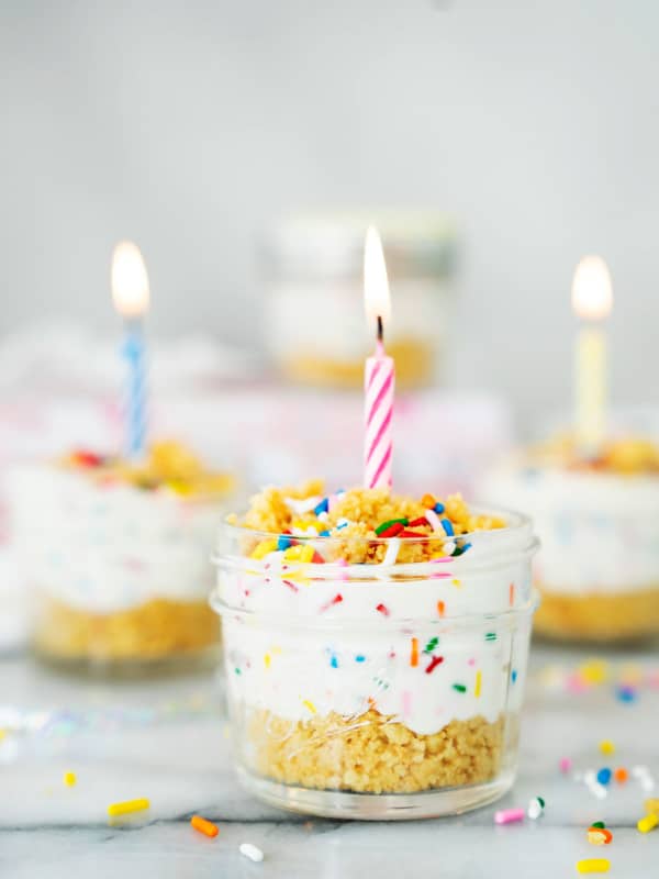 A small glass jar filled with layered cake and creamy frosting, topped with colorful sprinkles and a lit pink-striped candle. Similar jars with candles and sprinkles are blurred in the background.