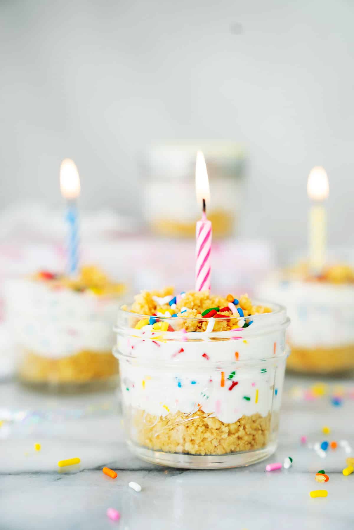 A small glass jar filled with layered cake and creamy frosting, topped with colorful sprinkles and a lit pink-striped candle. Similar jars with candles and sprinkles are blurred in the background.