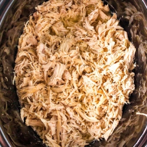 Shredded cooked chicken fills the inside of a black slow cooker, with the chicken appearing moist and tender, ready to be served or used in a recipe.