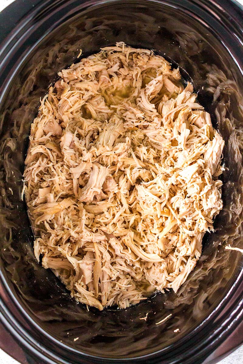 Shredded cooked chicken fills the inside of a black slow cooker, with the chicken appearing moist and tender, ready to be served or used in a recipe.