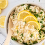 A bowl of cooked rice garnished with chopped parsley and three lemon slices, with a wooden spoon resting inside. Lemon slices and parsley are scattered on the white surface nearby.