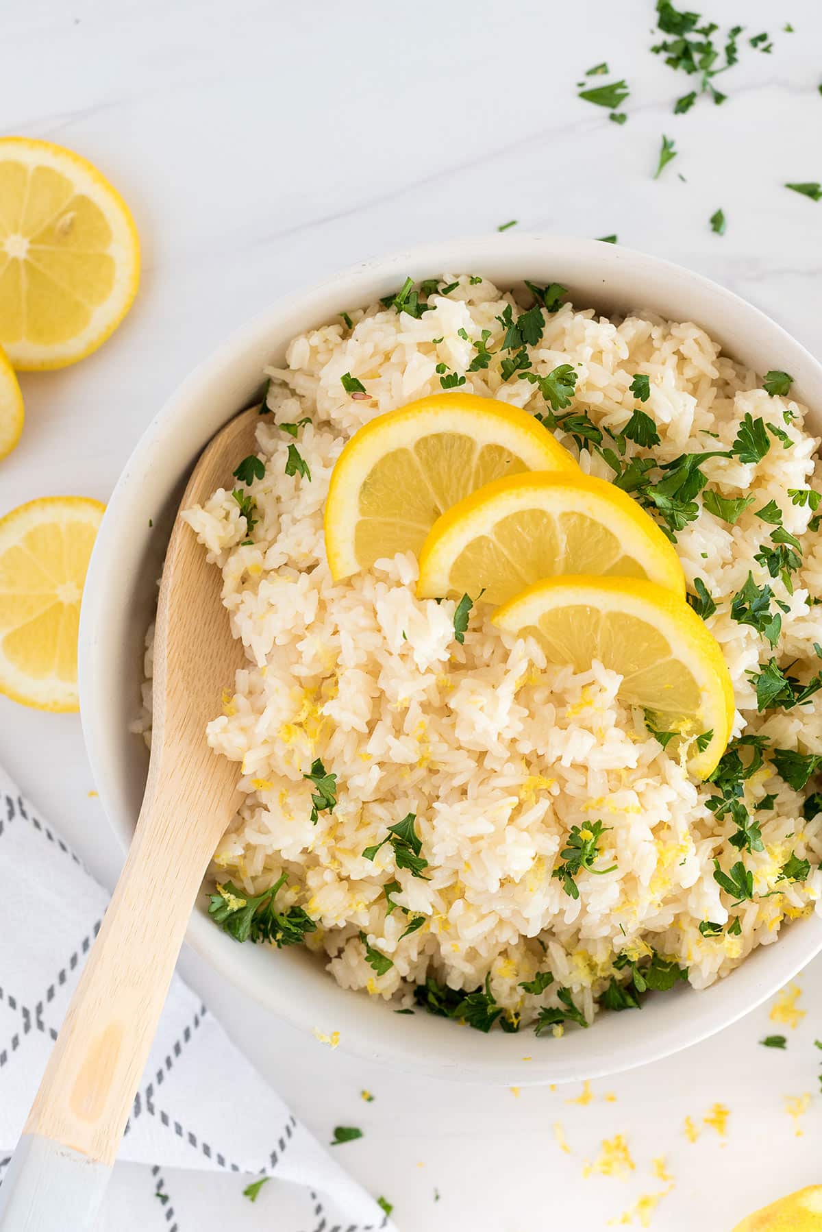A bowl of cooked rice garnished with chopped parsley and three lemon slices, with a wooden spoon resting inside. Lemon slices and parsley are scattered on the white surface nearby.