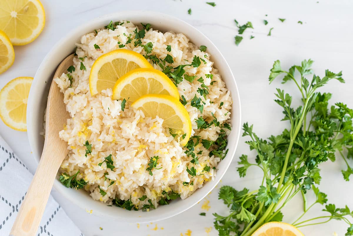 A bowl of rice garnished with lemon slices and chopped parsley, with a wooden spoon resting inside. Sprigs of fresh parsley and lemon slices are scattered on a white surface nearby.