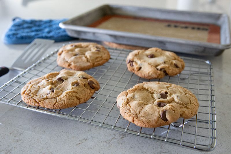 Four freshly baked chocolate chip cookies cool on a wire rack, with a baking tray lined with a silicone mat and a blue oven mitt in the background on a light-colored countertop.