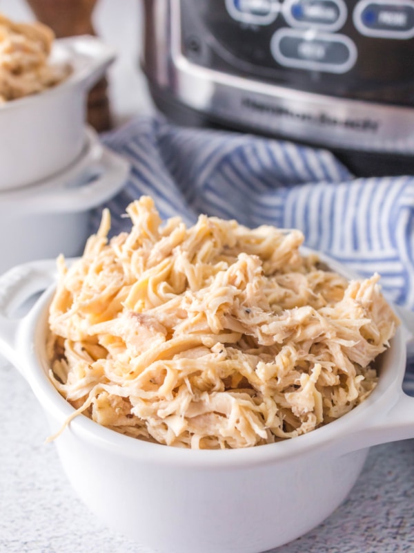 A white bowl filled with creamy shredded chicken sits on a light surface. A striped cloth is in the background along with a slow cooker and another bowl of shredded chicken.