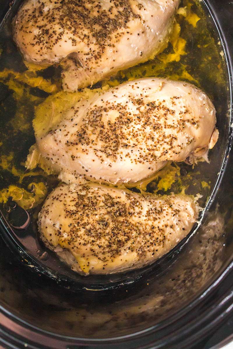 Three seasoned chicken breasts are cooking in a slow cooker, surrounded by juices. The chicken appears tender and moist, with visible black pepper and herbs sprinkled on top.