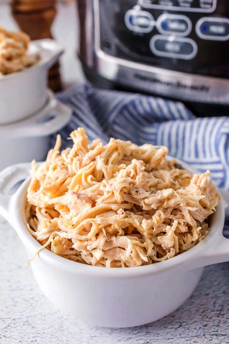 A white bowl filled with shredded chicken sits on a light surface, with a blue-striped cloth and a slow cooker in the background. The chicken appears tender and moist.