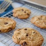 Four chocolate chip cookies cooling on a wire rack, with a baking tray and spatula in the background. The cookies are golden brown and chunky, sitting on a kitchen countertop.