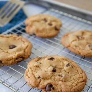 Four chocolate chip cookies cooling on a wire rack, with a baking tray and spatula in the background. The cookies are golden brown and chunky, sitting on a kitchen countertop.