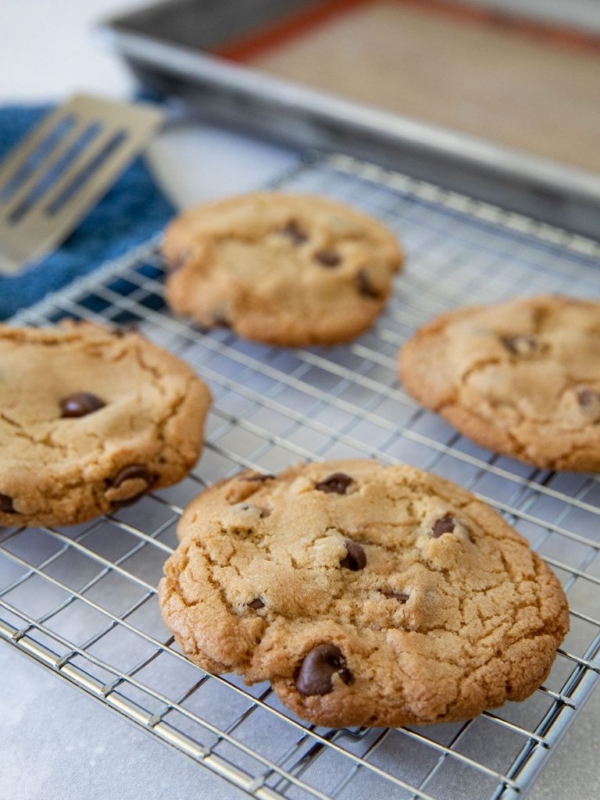 Four chocolate chip cookies cooling on a wire rack, with a baking tray and spatula in the background. The cookies are golden brown and chunky, sitting on a kitchen countertop.