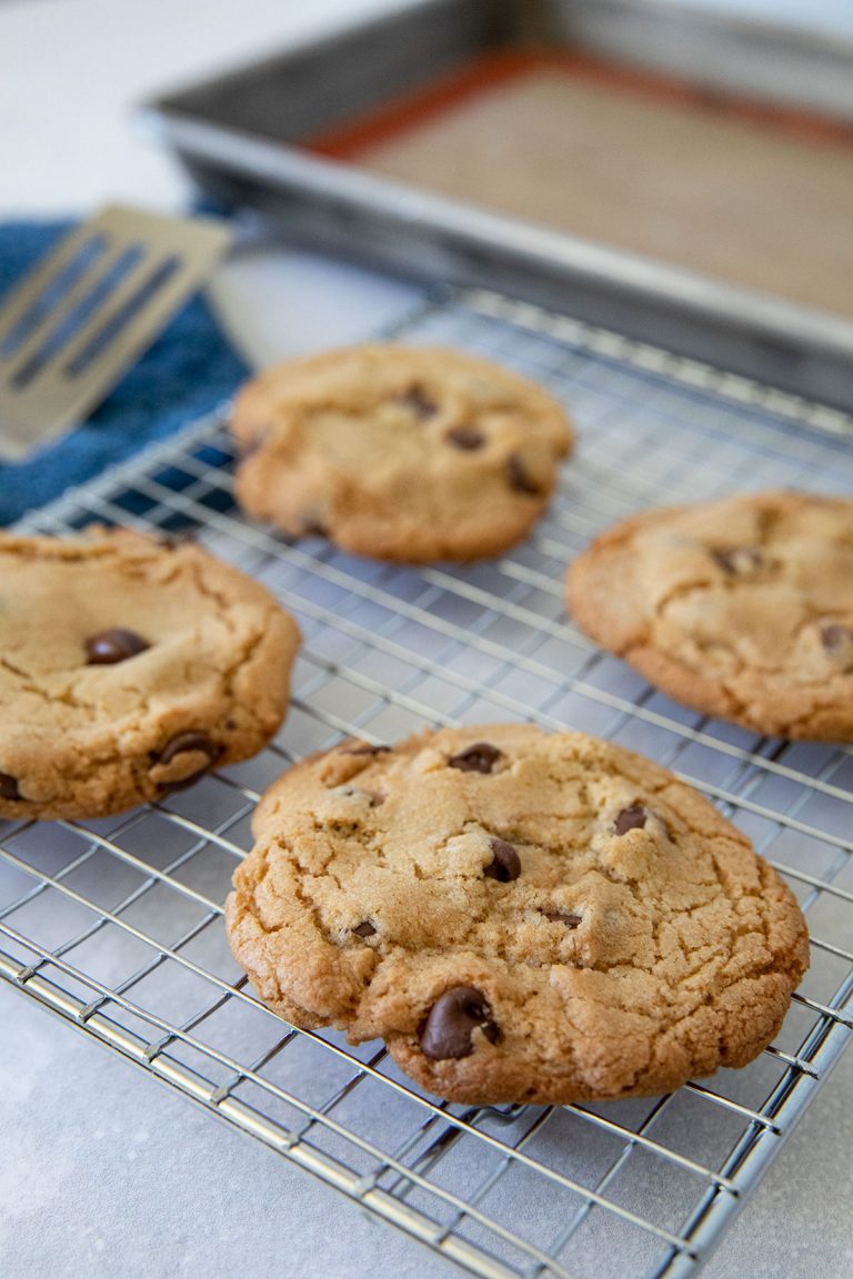 Four chocolate chip cookies cooling on a wire rack, with a baking tray and spatula in the background. The cookies are golden brown and chunky, sitting on a kitchen countertop.