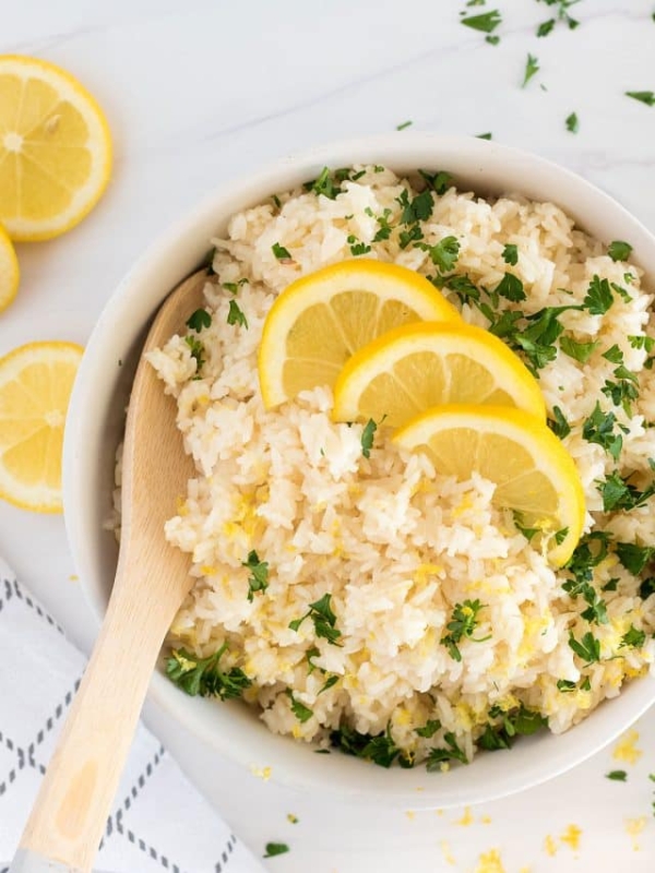 A white bowl filled with rice garnished with chopped parsley and slices of lemon, with a wooden spoon resting in the bowl. Lemon slices and parsley are scattered on the white surface nearby.