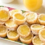 A plate of mini lemon tarts with golden yellow filling, sprinkled with powdered sugar. A small jar of lemon curd and a cup are in the blurred background.