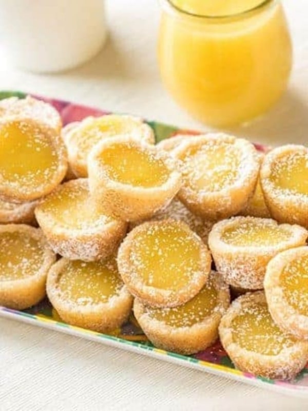 A plate of mini lemon tarts with golden yellow filling, sprinkled with powdered sugar. A small jar of lemon curd and a cup are in the blurred background.