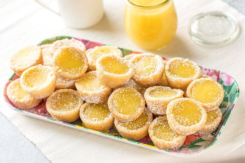 A colorful tray filled with mini lemon tartlets dusted with powdered sugar, with a glass jar of lemon curd and a milk jug in the background.