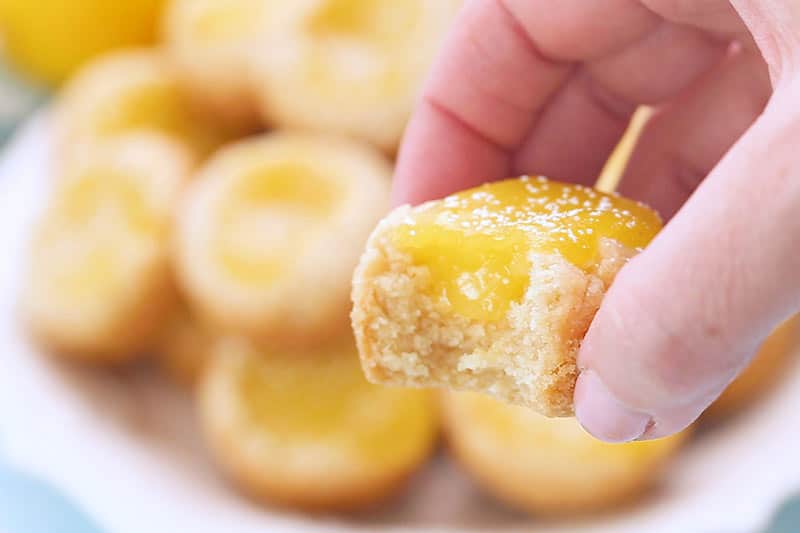 A hand holds a half-eaten lemon tart with yellow filling and powdered sugar, with more tarts blurred in the background.
