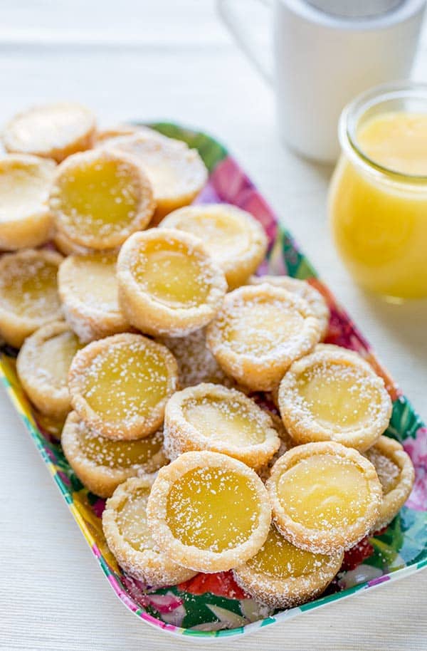 A platter of lemon curd tartlets dusted with powdered sugar, arranged in a pile, with a jar of lemon curd and a cup in the background.