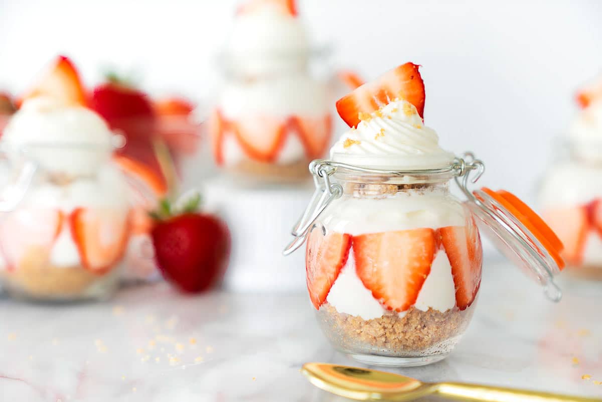 A small glass jar layered with graham crumbs, cream, and sliced strawberries, topped with whipped cream and a strawberry slice. More jars and strawberries are blurred in the background. A gold spoon sits nearby.