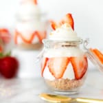 A glass jar with layered cheesecake, sliced strawberries, and whipped cream on top. A gold spoon and more strawberries are in the background, with another dessert jar slightly blurred behind.