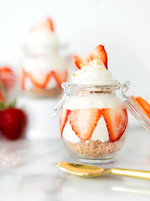 A glass jar with layered cheesecake, sliced strawberries, and whipped cream on top. A gold spoon and more strawberries are in the background, with another dessert jar slightly blurred behind.