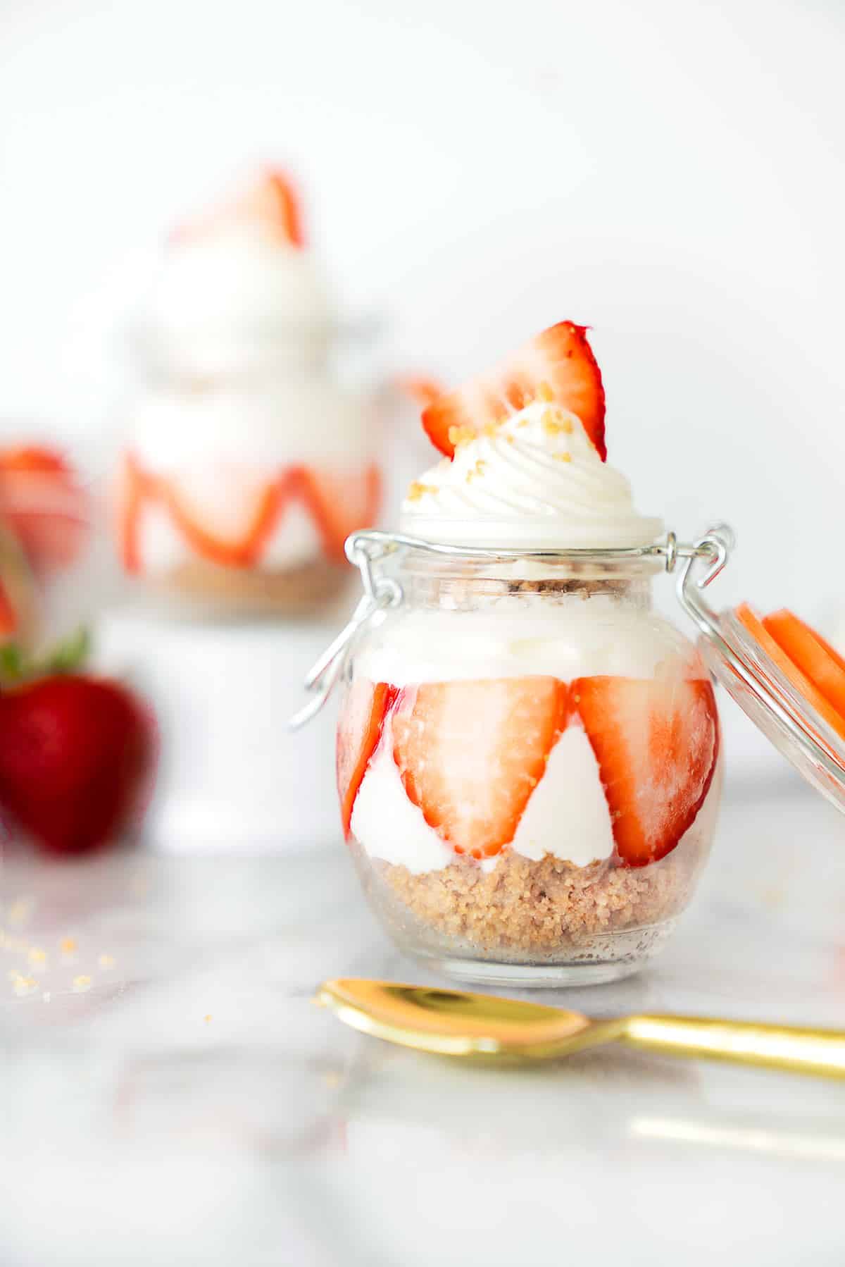 A glass jar with layered cheesecake, sliced strawberries, and whipped cream on top. A gold spoon and more strawberries are in the background, with another dessert jar slightly blurred behind.