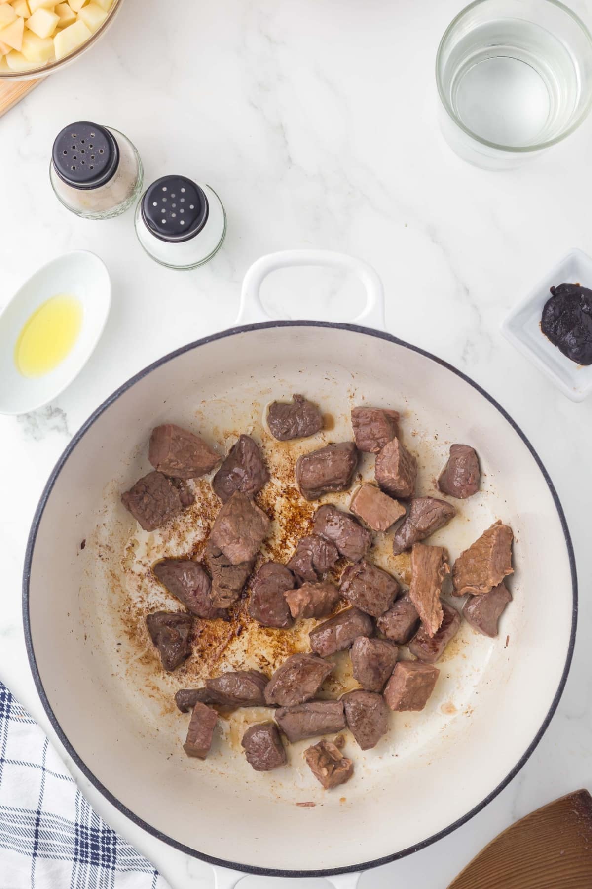 Chunks of browned beef cook in a white pot on a marble counter, surrounded by salt, pepper, a bowl of water, olive oil, diced potatoes, a prune, and a towel.