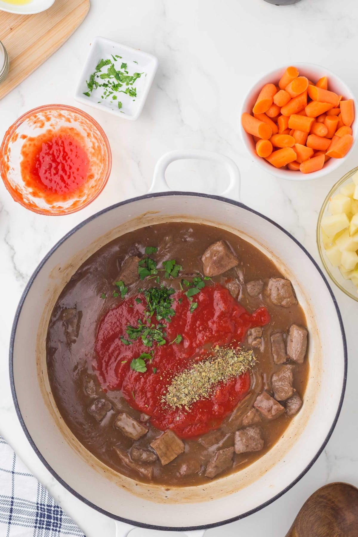Beef stew in a white pot with tomato sauce, herbs, and parsley on top. Surrounding the pot are bowls of chopped carrots, potatoes, and parsley, plus a bowl with tomato sauce on a white marble surface.