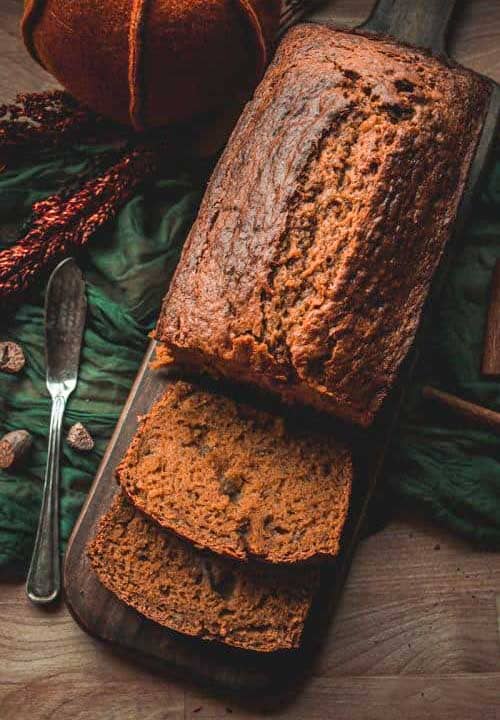 A loaf of pumpkin bread with two slices cut sits on a dark wooden board, surrounded by a butter knife, pumpkin, and spices on a rustic table.
