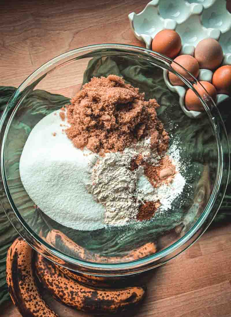A glass bowl with sugar, brown sugar, flour, and spices sits on a wooden surface next to ripe bananas and a white dish holding several eggs.