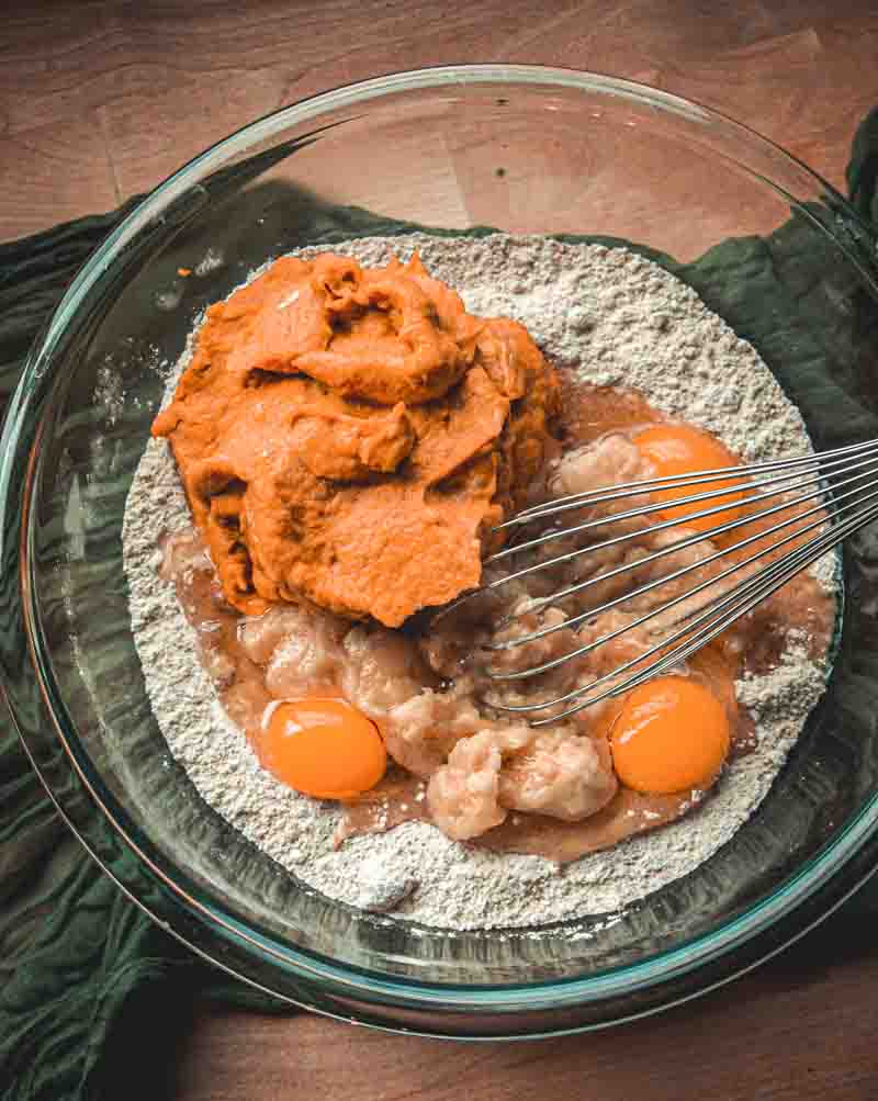 A glass bowl holds flour, eggs, mashed bananas, and a scoop of pumpkin puree being mixed with a whisk on a wooden surface.