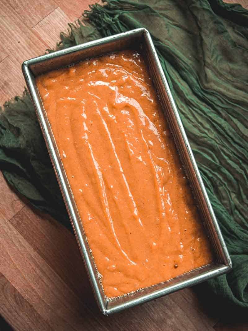 A metal loaf pan filled with orange batter sits on a wooden surface, next to a dark green textured cloth.