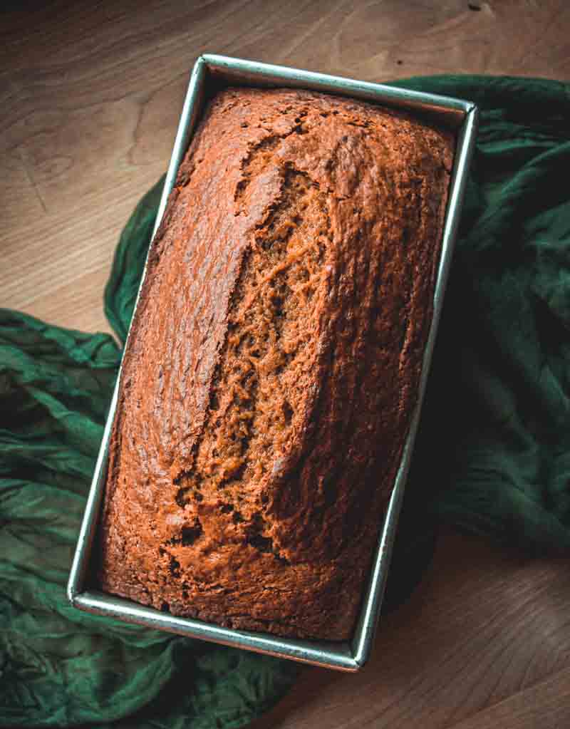 A golden-brown loaf of banana bread sits in a rectangular metal pan on a wooden surface, next to a dark green cloth.