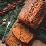 Sliced loaf of brown bread on a wooden board, with two slices cut. A butter knife and green cloth are nearby, along with some spices and decorative dried plants.