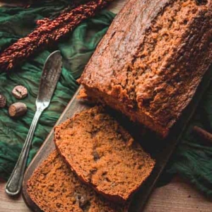 Sliced loaf of brown bread on a wooden board, with two slices cut. A butter knife and green cloth are nearby, along with some spices and decorative dried plants.