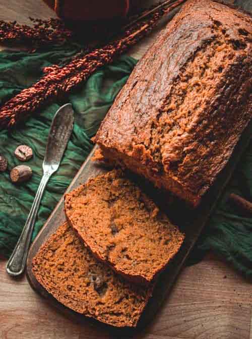 Sliced loaf of brown bread on a wooden board, with two slices cut. A butter knife and green cloth are nearby, along with some spices and decorative dried plants.